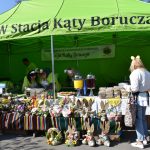 Na zdjęciu widać zielony namiot z napisem KGiGW Stacja Kąty Borucza. Przed nim stół z kolorowymi dekoracjami wielkanocnymi i jedzeniem. W tle trzy osoby w jasnych ubraniach przygotowują potrawy.