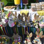 Na kolorowym straganie widoczne są ręcznie robione dekoracje wielkanocne. Słomiane króliczki mają duże uszy i ozdobne elementy. Tło to biała zasłona i różnokolorowe obrusy.