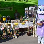 Stoisko targowe z zielonym namiotem, świąteczne dekoracje i napoje. Maskotka królika w liliowej sukni trzyma koszyk.