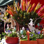 Na zdjęciu kolorowe kosze wielkanocne zdobią stół, otoczone kwiatami i pisankami. Duży bukiet kolorowych liści, tło to ciemny namiot. Dekoracje są żywe i świąteczne.