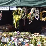 Kolorowy stragan z wieńcami wielkanocnymi, bogato zdobionymi kwiatami i wstążkami. Na stole ozdoby i pudełka z ciastami. Zielony namiot z napisem Rada Rodziców Dobre.