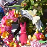 Kolorowy wieniec z kwiatami, jajkami i motylkiem; różowy królik na tle liści. Pastelowe i żywe barwy na białym tle.