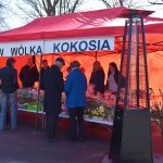 Na zdjęciu widać czerwony namiot targowy z napisem „KGW Wólka Kokosia”. Pod namiotem ludzie stoją przy stoliku pełnym produktów, takich jak kwiaty i jedzenie. W tle są drzewa i znak „Gra terenowa”.