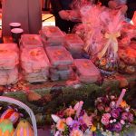 Stół z kolorowymi dekoracjami wielkanocnymi i jedzeniem. W tle pomarańczowy namiot, na stole pudełka z jedzeniem i koszyk z jajkami. Obok kwiatowe kompozycje z fioletowymi i żółtymi tulipanami.