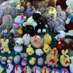 Kolorowe, ręcznie robione maskotki z włóczki na zielonym tle. Przedstawiają zwierzęta takie jak króliki, owce i kurczaki. Wielobarwna i różnorodna kolekcja.