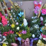 Barwne palmy wielkanocne w pudełku z zielenią, kwiatami i ozdobami. Dominują czerwone, żółte i fioletowe akcenty. Tło tekturowe.