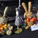 Na stole znajduje się grupa dekoracyjnych zajęcy z siana i włóczki. Mają kolorowe kokardy i rekwizyty jak marchewki. Tło jest ciemne, a na stole widnieje kartka z napisem II kategoria Dzieci 11-10 lat.