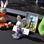 Kilka dekoracyjnych zajączków wielkanocnych na czarnym tle. Jeden z wiązki, drugi biały na szydełku, a trzeci zielony z puszystym futerkiem. Obok nich kolorowe kwiaty i pisanki.