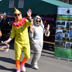 Na zdjęciu dwie osoby w kolorowych kostiumach świętują na świeżym powietrzu. W tle zielony namiot, a po prawej banner promocyjny. Pogodny, słoneczny dzień z uczestnikami wydarzenia w tle.