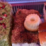 Różnorodne ciasta na tacy, wśród nich zielony deser z pestkami granatu. Tło różowe, ciasta różnobarwne z kruszonką.