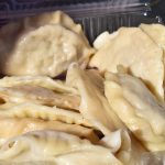 Na zdjęciu widać pierogi w przezroczystym pojemniku. Mają złocisty kolor i są ułożone na siebie. Tło jest rozmyte, w odcieniach zieleni.