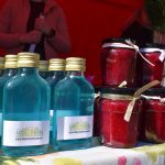 Na zdjęciu kolorowe napoje w niebieskich butelkach i czerwone przetwory w słoikach na tle czerwonego namiotu. Tkanina dekoracyjna na stole w kwiaty. W tle widoczne osoby.
