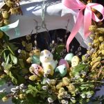 Wieniec z wierzby z kolorowymi pisankami i pluszowym kurczakiem. Zielone liście i kokardy na białym tle. Pastelowe ozdoby.