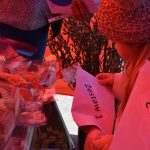 Osoba trzyma zestaw pod nazwą Zestaw 1 przy stoisku z upominkami. Ciepłe kolory tła tworzą świąteczną atmosferę. Rośliny i tulipan w tle dodają wiosennego klimatu.