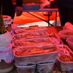 Na zdjęciu widać stoisko z żywnością na targu. Produkty są w plastikowych i metalowych pojemnikach z folią. Tło pomarańczowe, dodające ciepłej barwy.