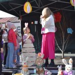 Grupa dzieci na scenie na zewnątrz, ozdobiona kolorowymi ozdobami. Kobieta w różowej spódnicy rozmawia z dziećmi. W tle widać dekoracje wielkanocne z motywem zajączka.