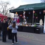 Scena z zielonym namiotem na festynie wielkanocnym. Dzieci stoją przed stołem z ozdobami, obok namiotu z czerwonym daszkiem. Kolorowe koła na ziemi.