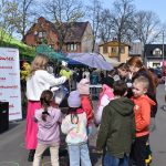 Na zdjęciu jest grupa dzieci i dorosłych, a w tle namioty z napisami. Tło to zielone i różowe namioty oraz budynki. Obok jest zielone drzewo bez liści na niebieskim tle nieba.