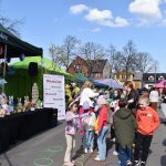 Na zdjęciu widać zielone namioty z dekoracjami i grupę dzieci na placu. Jasne niebo i wiosenna atmosfera. Tło to zabudowania i drzewa bez liści.