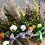 Bukiety wielkanocne na stole z jasnego drewna. Dominują zielone gałązki, białe, pomarańczowe kwiaty i kolorowe wstążki. W tle asfaltowa powierzchnia.