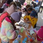 Dzieci kolorują ozdoby przy drewnianym stole, używając pisaków. W tle widać kolorowe namioty i dorosłych. Jest jasno i pogodnie.