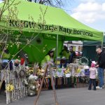 Zielony namiot z napisem, stoisko z ozdobami wielkanocnymi. W tle drzewa, ludzie przeglądają kolorowe produkty.