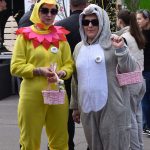 Dwóch ludzi w kostiumach zwierząt trzyma różowe koszyki. Tło z gałązkami i zielonym namiotem. Kolorowe i wesołe otoczenie.