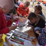 Dzieci przy stole dekorują kolorowe papierowe wycinanki. W tle różowy namiot na tle budynków. Atmosfera jest twórcza i wesoła.