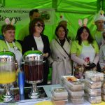 Grupa osób ubrana w stroje świąteczne stoi pod zielonym namiotem. Na stole znajdują się soki i ciasta w plastikowych opakowaniach. W tle jest baner z napisem promującym wydarzenie w Kątach Borucza.