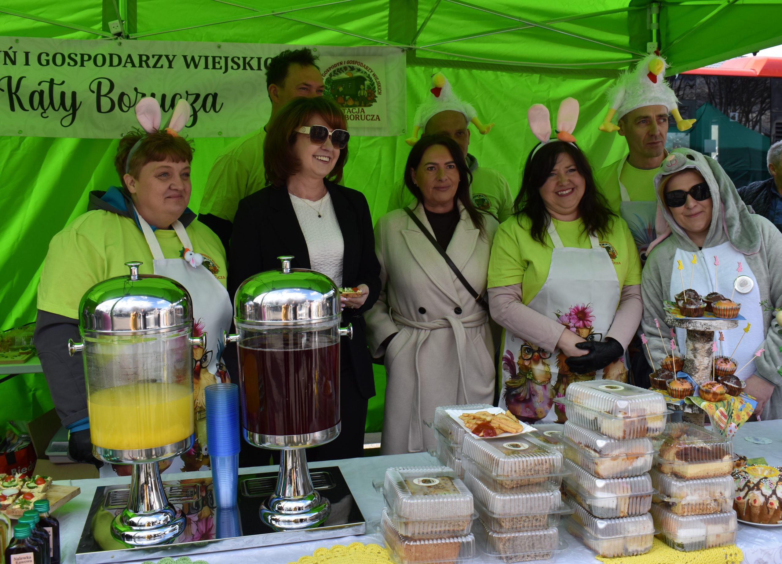 Grupa osób ubrana w stroje świąteczne stoi pod zielonym namiotem. Na stole znajdują się soki i ciasta w plastikowych opakowaniach. W tle jest baner z napisem promującym wydarzenie w Kątach Borucza.