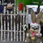 Pleciony zając z trawy o dużych oczach trzyma gałązki. Obok wiklinowy płot z brzozy, dekoracje i kolorowe tło. W tle stół z kwiatami.