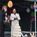 Na zdjęciu dwie kobiety na scenie, jedna trzyma mikrofon. Tło zdobią kolorowe dekoracje papierowe, w tym jajko. Na przodzie są dekoracyjne zające.