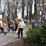 Na zdjęciu grupa ludzi w parku w jasnych ubraniach. W tle widoczny ceglany kościół. Drzewa i dekoracje wielkanocne na krzewach.