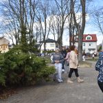 Grupa ludzi spaceruje po parku z iglakami ozdobionymi pisankami. W tle są kolorowe budynki. Scena ma pogodny, wiosenny charakter.