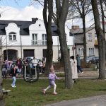 Plac z drzewami, po którym dzieci w kolorowych ubraniach bawią się i biegają. W tle białe i szare budynki, kilka osób spaceruje. Atmosfera wiosenna.