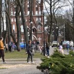 Na zdjęciu widzimy ludzi spacerujących po parku z ceglaną, gotycką kaplicą w tle. Drzewa są bez liści, a na pierwszym planie widać zielony krzew ozdobiony kolorowymi jajkami. Pogoda jest pochmurna, co sugeruje chłodniejs