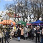 Targ na świeżym powietrzu z kolorowymi namiotami. W tle drzewa i budynki. Ludzie spacerują po placu w pogodny dzień.