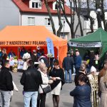 Na zdjęciu jest targ na świeżym powietrzu z pomarańczowym i zielonym namiotem. Ludzie w kurtkach stoją i rozmawiają. W tle są drzewa i budynki.