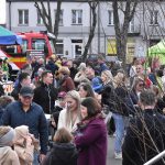 Na zdjęciu widoczna jest duża grupa ludzi podczas festynu na świeżym powietrzu. W tle stoi czerwony wóz strażacki, a obok są kolorowe namioty. Uczestnicy ubrani są w różnokolorowe kurtki, co tworzy żywą i rados