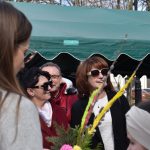 Na zdjęciu grupa ludzi na tle zielonego namiotu. Osoby są uśmiechnięte, jedna trzyma kolorowe gałązki. W tle widać drzewa.