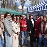 Grupa ludzi stoi razem na tle białego i niebieskiego namiotu w parku. Ubrani są w płaszcze i kurtki w odcieniach beżu i czerwieni. Tło stanowią drzewa bez liści i błękitne niebo z chmurami.