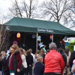 Na zdjęciu widać zielony namiot z napisem Gmina Dobre oraz tłum ludzi w kolorowych ubraniach. Na tle jasnego nieba stoją nagie drzewa. Scena udekorowana jest kolorowymi ozdobami.