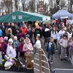 Ludzie na festynie w parku, wokół straganów z namiotami. Dzieci w kolorowych kurtkach, w tle zielony namiot i drzewa. Dekoracje w kształcie zajączków na stole.