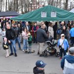 Na zdjęciu są ludzie w różnym wieku na targu z kramami. Dominują kolory: zielony namiot, kolorowe ubrania i ciemne kurtki. W tle widać drzewa i budynki.