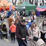 Na zdjęciu widać tłum ludzi przy stoiskach na zewnątrz. Namioty są w kolorach pomarańczowym, zielonym i niebieskim. Tło uzupełniają drzewa i budynki.