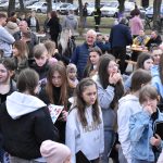 Na zdjęciu tłum ludzi na zewnątrz w odzieży zimowej. W tle widać samochody i drzewa. Osoby różnego wieku stoją blisko siebie.