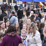 Tłum ludzi na zewnątrz, głównie dzieci i kobiety. W tle widać stragany i wóz strażacki. Kolorowe ubrania, wiosenna aura.