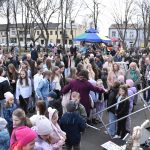 Na zdjęciu widać tłum osób na wydarzeniu plenerowym, w tym dzieci i dorosłych. W tle widać kolorowe namioty i drzewa. Ludzie ubrani są w różnorodne ubrania, a atmosfera jest wesoła.