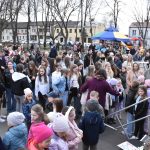 Na zdjęciu widoczny jest tłum ludzi na zewnątrz, otoczony drzewami i budynkami. W tle są kolorowe stragany i namioty z niebieskim i żółtym daszkiem. Uczestnicy, w różnorodnych strojach, wydają się brać