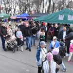 Ludzie spacerują po jarmarku z kolorowymi namiotami. Jest pochmurno, ale atmosfera jest radosna. W tle widoczne są wysokie drzewa i budynki.